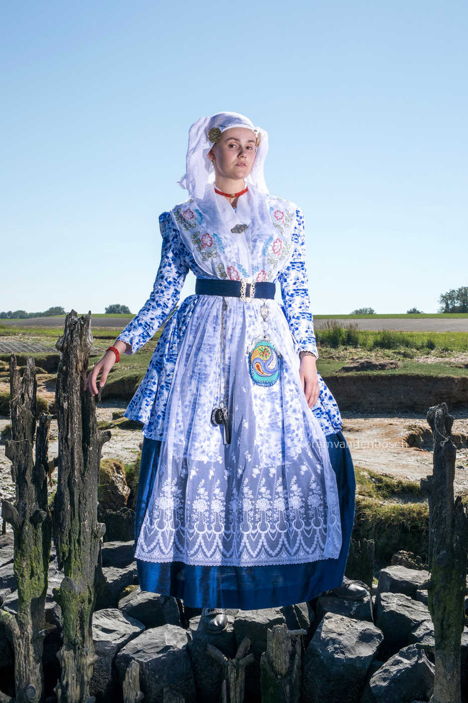 Linde in blauw-witte traditionele Friese dracht staand tussen oude paalhoofden aan zee met een speelse blik. Fotografie Rem van den Bosch 