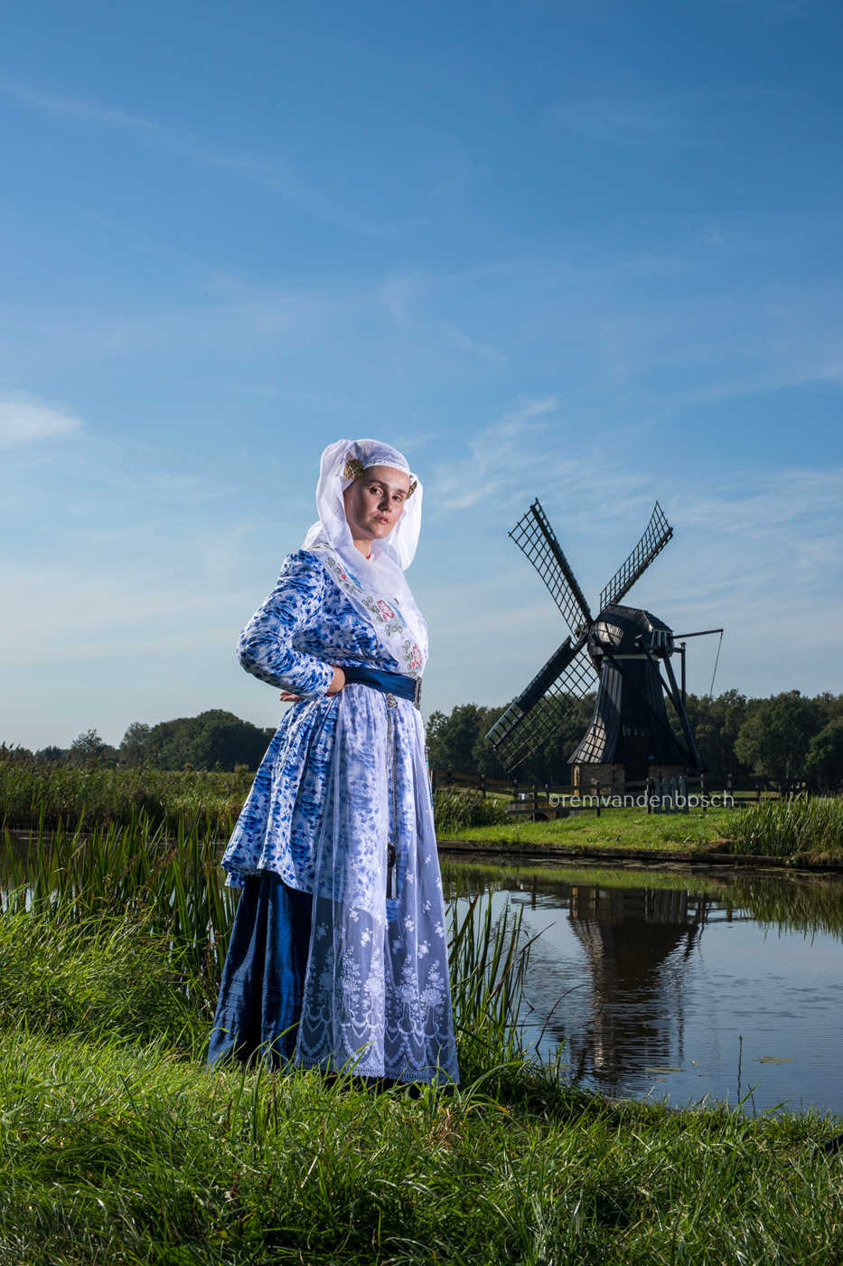 Linde in blauw-witte Friese dracht in het gras aan het water met een oude houten windmolen op de achtergrond. Fotografie Ren van den Bosch 