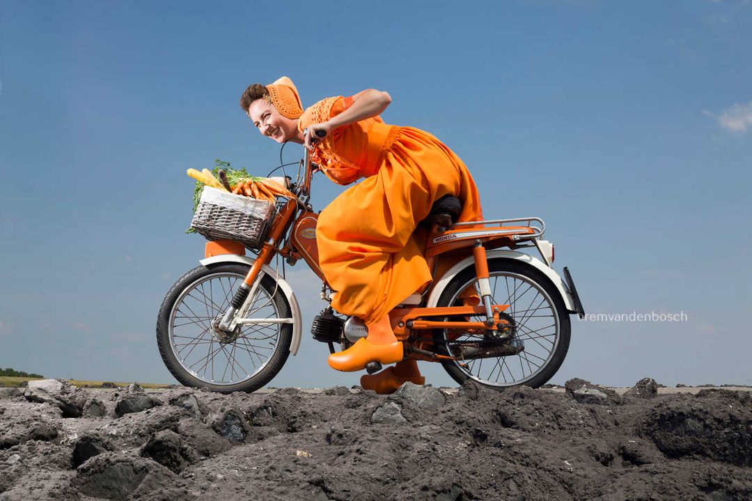 Liggend fotobeeld van Ruby in oranje Walcherse dracht op een brommer met een mand vol wortels op de dijk, Rem van den Bosch
