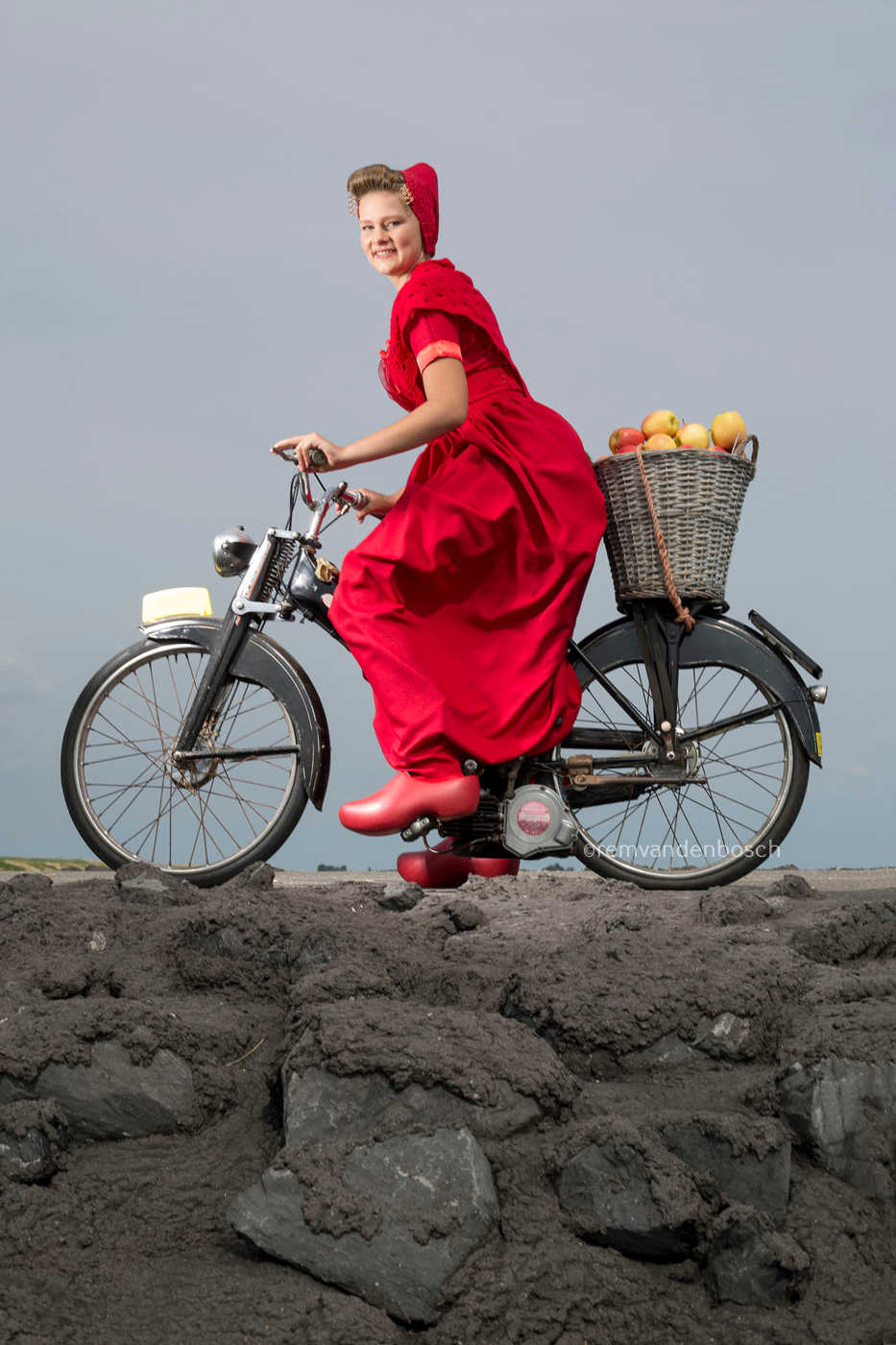 Staand portret van Phoebe in moderne rode Walcherse dracht op de dijk met een brommer en een mand vol appels