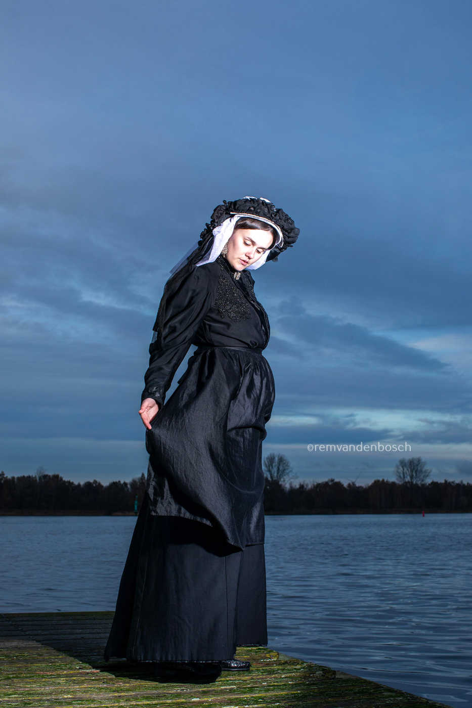 Linde in traditionele Limburgse dracht op een houten steiger aan het water, ingetogen naar de vloer kijkend. Fotografie Rem van den Bosch 
