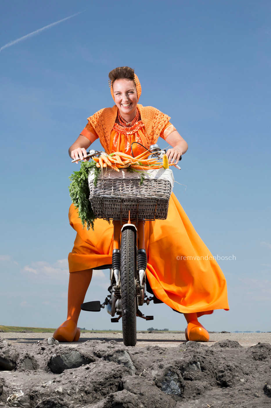 Ruby in oranje Walcherse dracht op een brommer met een mand vol wortels op de dijk , Zeeuwse wandkunst, Rem van den Bosch 
