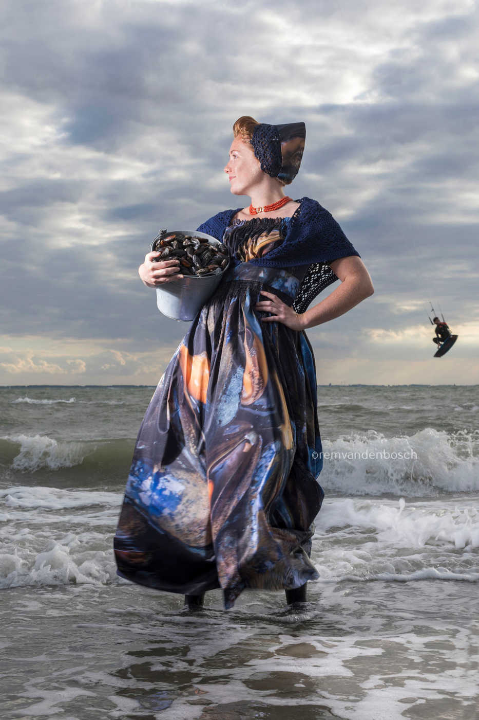 Sharon in moderne Walcherse dracht met mosselprint, staand in de zee met een emmer mosselen en een kitesurfer op de achtergrond
