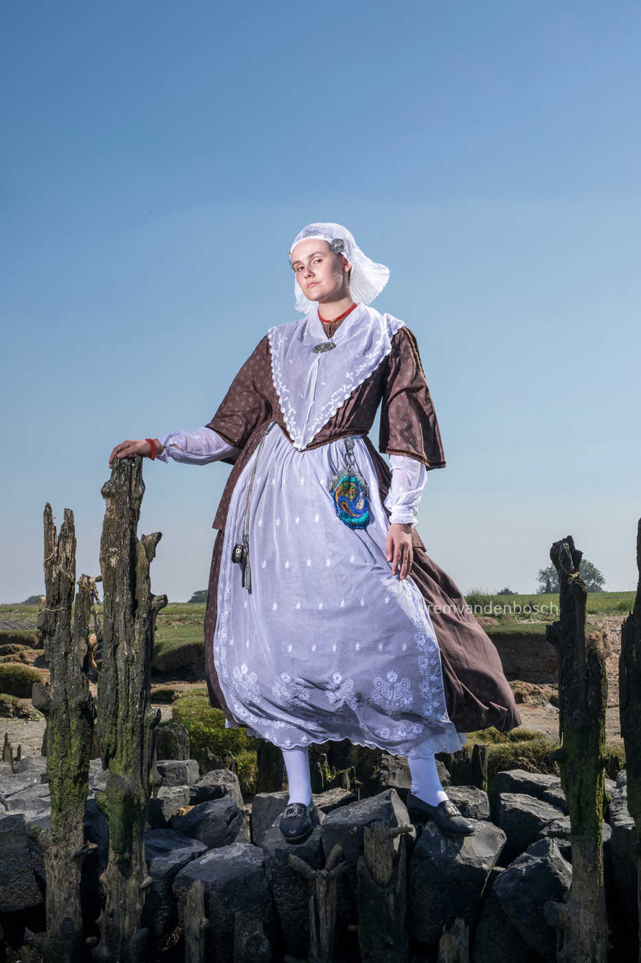 Linde in traditionele Friese dracht staand tussen oude paalhoofden aan zee met een speelse blik